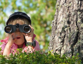 Junges Mädchen sitzt in der Natur neben einem Baum und schaut mit einem Fernglas in die Ferne